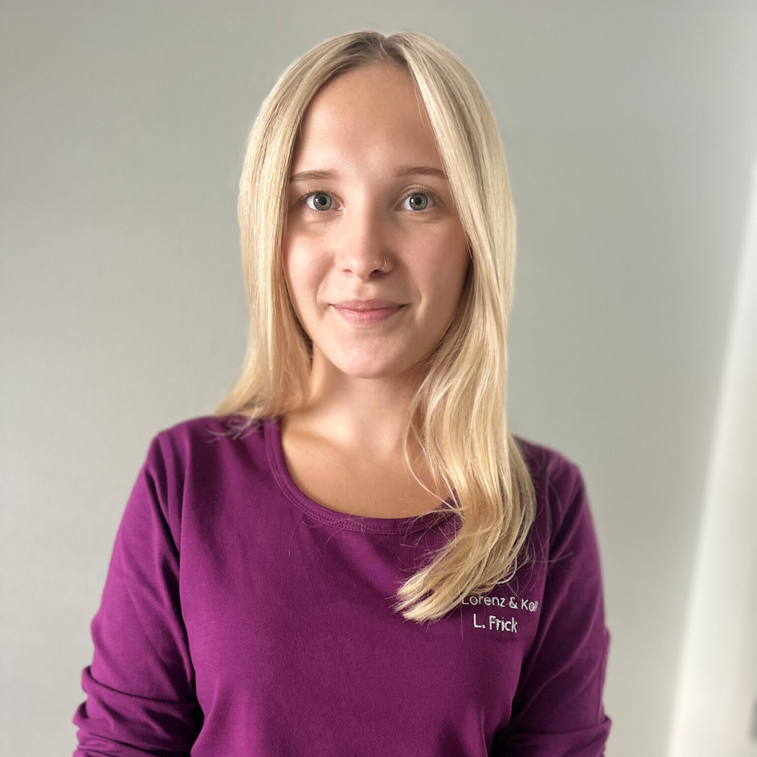 Portrait von Lilly Frick Junge blonde Frau mit offenen Haaren und Nasenpiercing, trägt ein violettes Oberteil mit der Aufschrift „Dr. Lorenz & Koll. – L. Frick“, steht lächelnd vor neutralem Hintergrund.